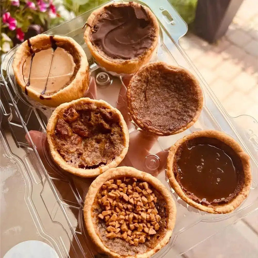 A close-up of five clamshells of tarts in a display, featuring Maple Butter, Pecan Honey, and Sea Salt Chocolate flavors arranged on a wooden tray.